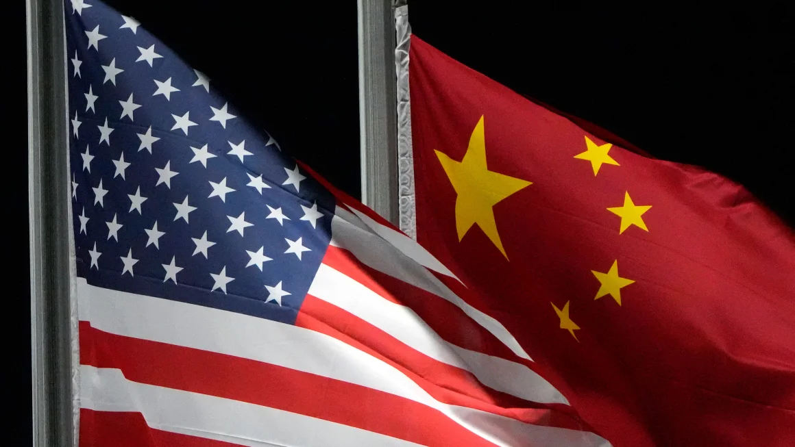 Dos banderas ondeando juntas: la bandera de los Estados Unidos a la izquierda y la bandera de China a la derecha. Ambas banderas están parcialmente superpuestas, lo que simboliza la relación entre las dos naciones. La bandera estadounidense es fácilmente reconocible por sus franjas rojas y blancas con un campo azul lleno de estrellas, mientras que la bandera china es roja con una gran estrella amarilla acompañada de cuatro estrellas más pequeñas. El fondo es oscuro, destacando los colores vivos de las banderas.