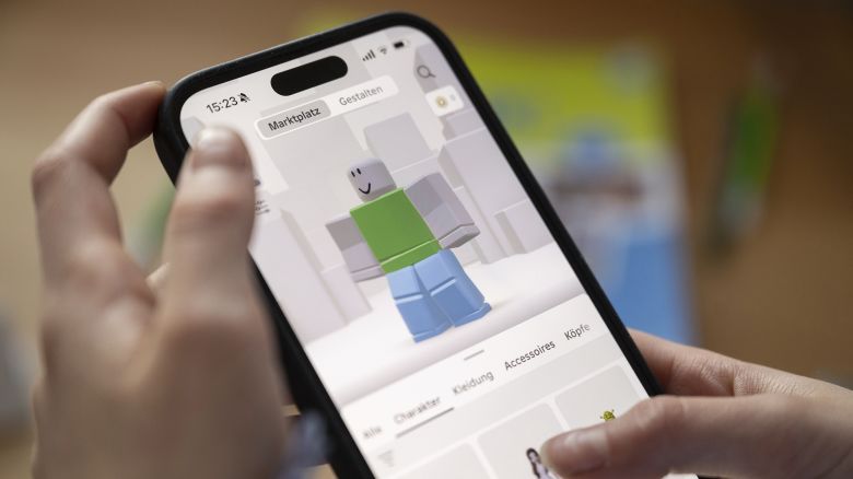 A girl operates a smartphone on which the Roblox app can be seen.