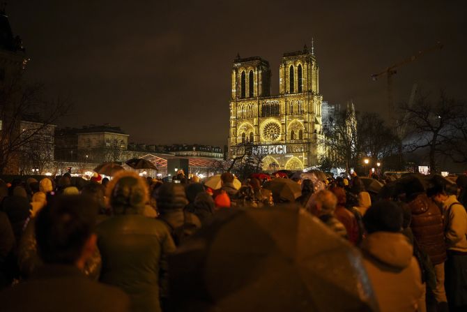 Spectators gather outside Notre Dame as it is lit with the words for "Thank you" in multiple languages.