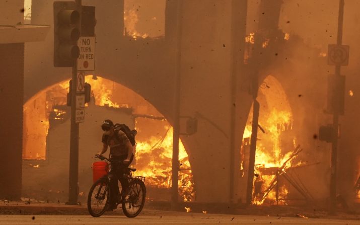 A cyclist pedals past a burning structure in Altadena.