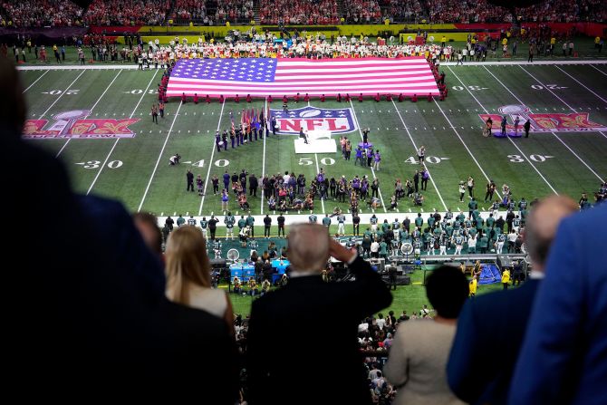 President Donald Trump, in the foreground, salutes as Jon Batiste performs the National Anthem before the game.