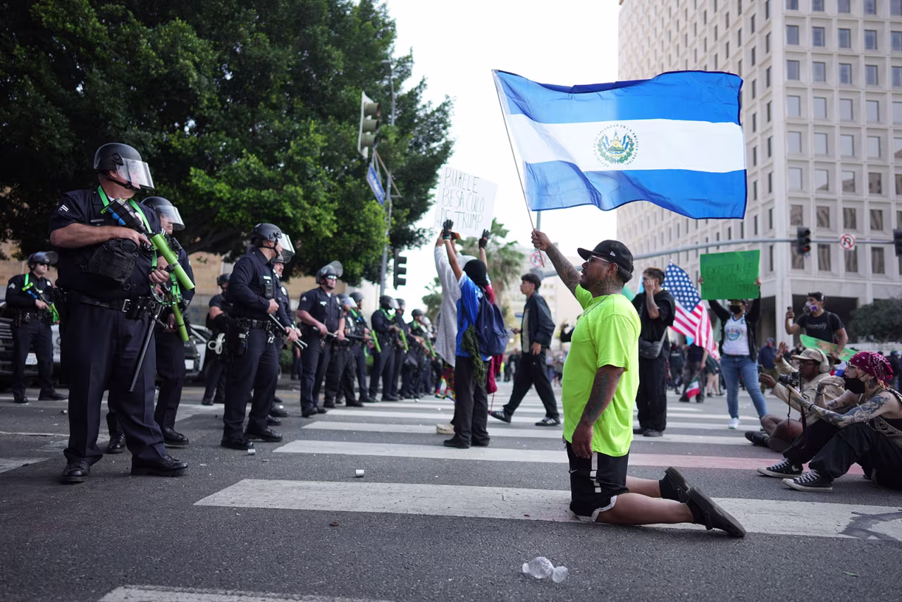 A protester kneels waving a National flag from El Salvador in front of a line of law enforcement officers near the Federal Building in downtown Los Angeles on Monday.