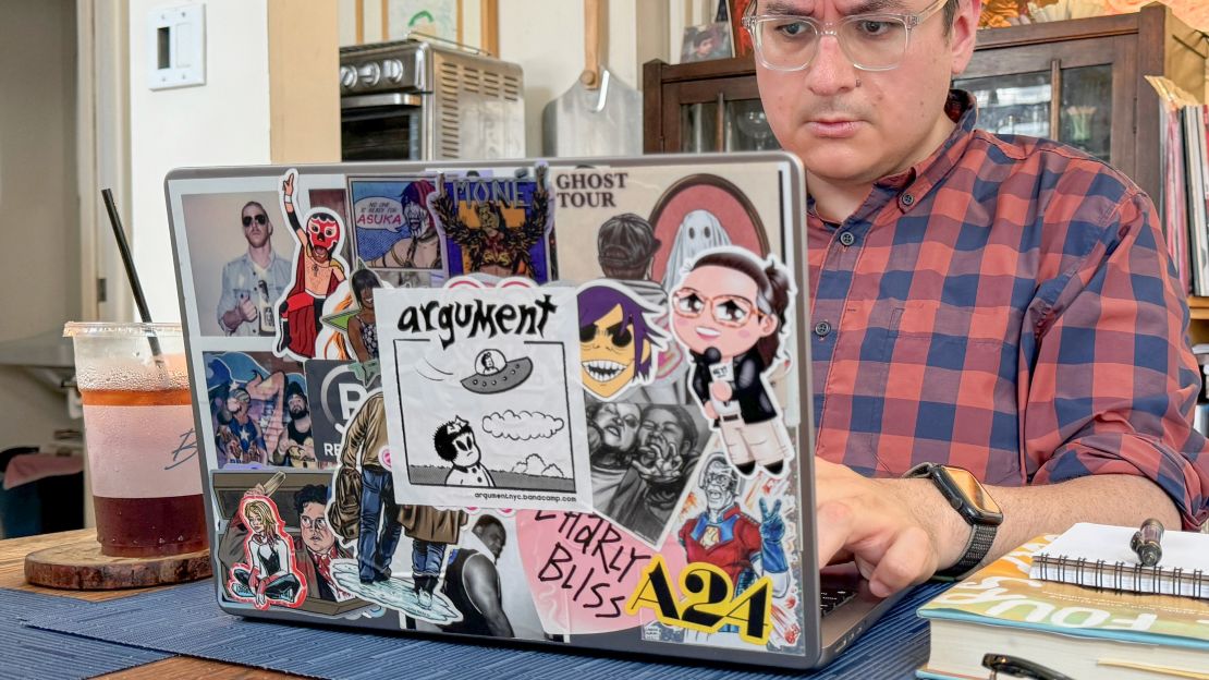 Henry is typing on a 14-inch MacBook Pro we see from behind, its lid obscured by layers of stickers.