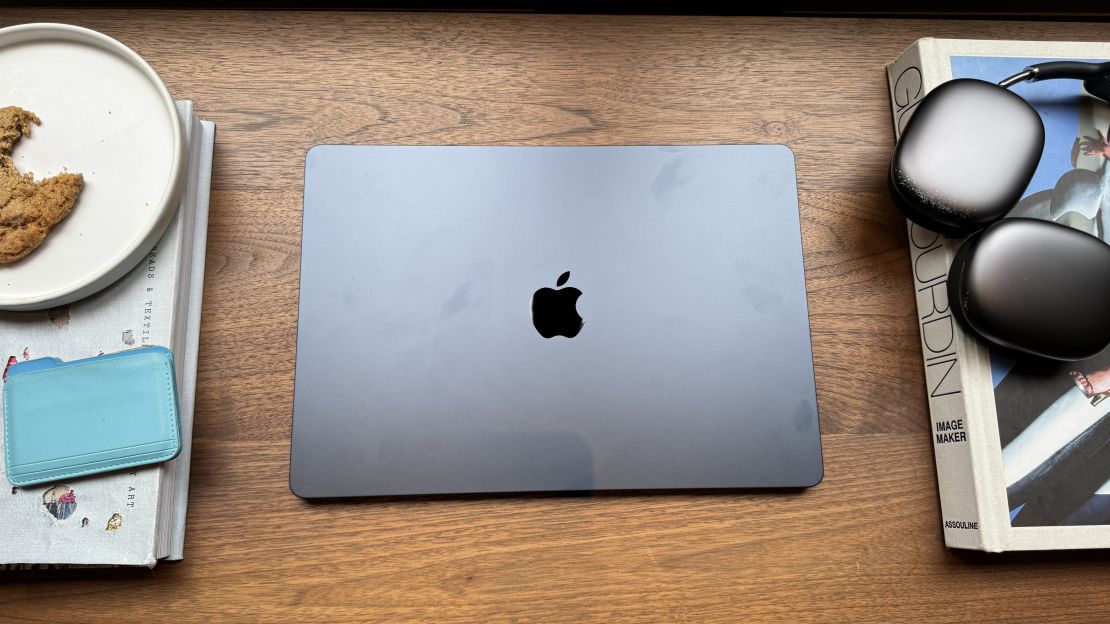 A Midnight Apple MacBook Air M3 with minor fingerprint smudges, on a table surrounded by a cookie, a blue wallet, books and AirPods Max