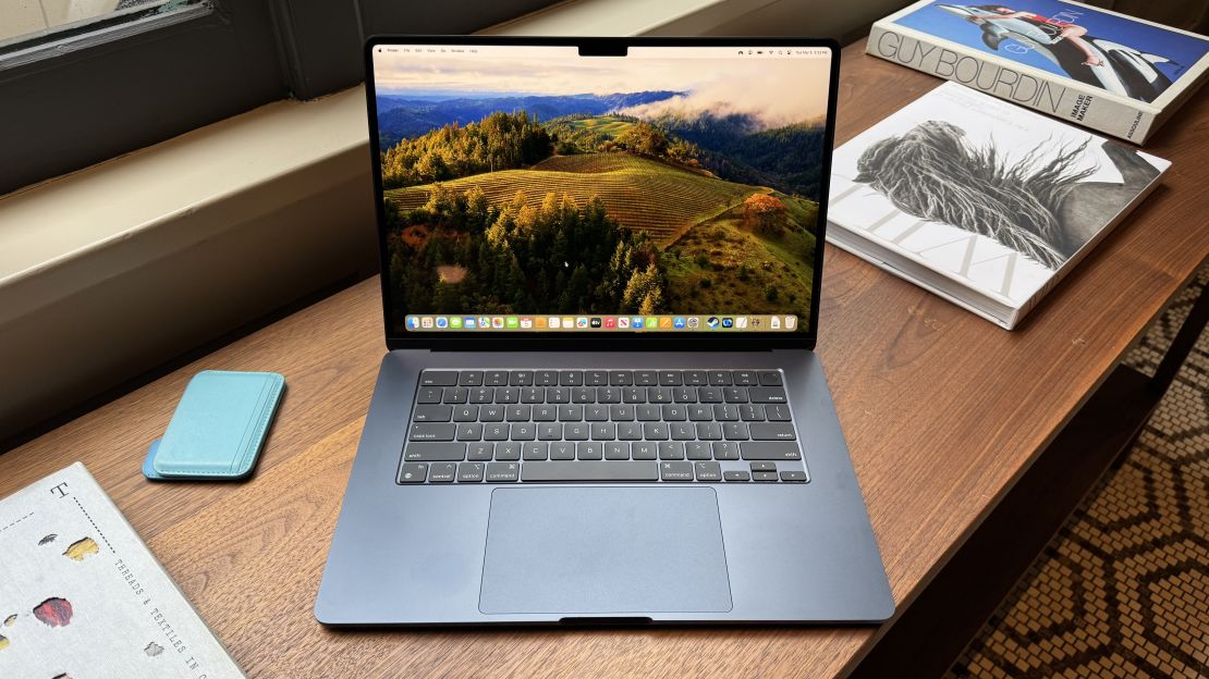 The 15-inch Midnight Blue Apple MacBook Air M3 has a scenic vista on its wallpaper, and sits open on a table with books on its sides.