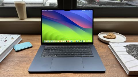 A MacBook Air with M3 (15-inch, Midnight blue) is open on a table, surrounded by a blue wallet, a coffee, a cookie and books.