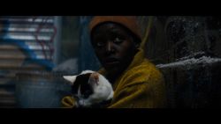 Lupita Nyong'o sits in an alley holding 'Frodo' the cat in a scene from 'A Quiet Place: Day One.' Nyong'o is wearing a yellow sweater and an orange wool cap.