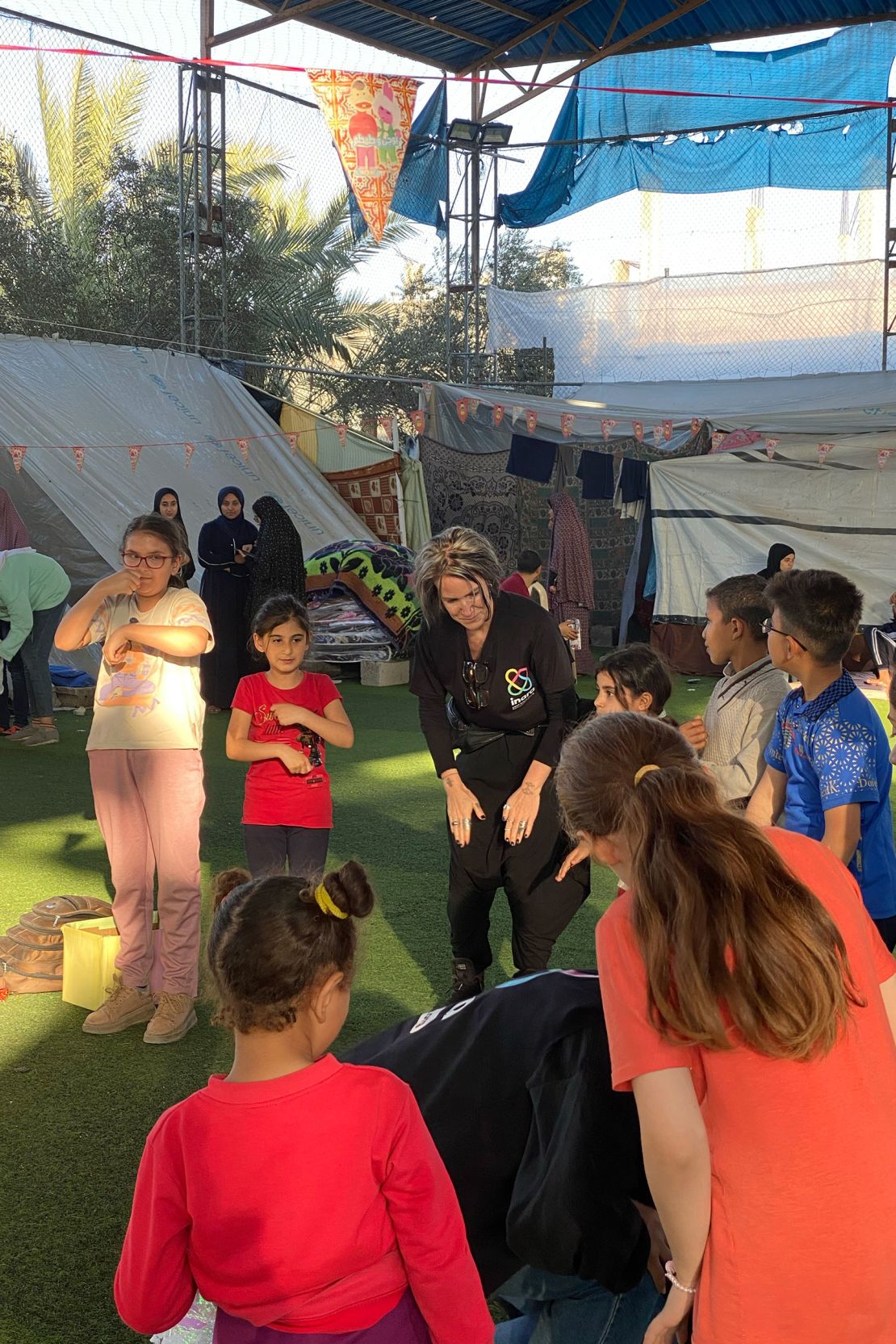 A woman claps her hands to her thighs, amidst a group of children. 