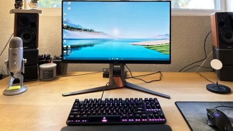 An Asus ROG Swift PG259QNR gaming monitor on a wooden desk, with keyboard, mouse, speakers and monitor visible on the desktop, backlit by an open window on the wall above the desk.