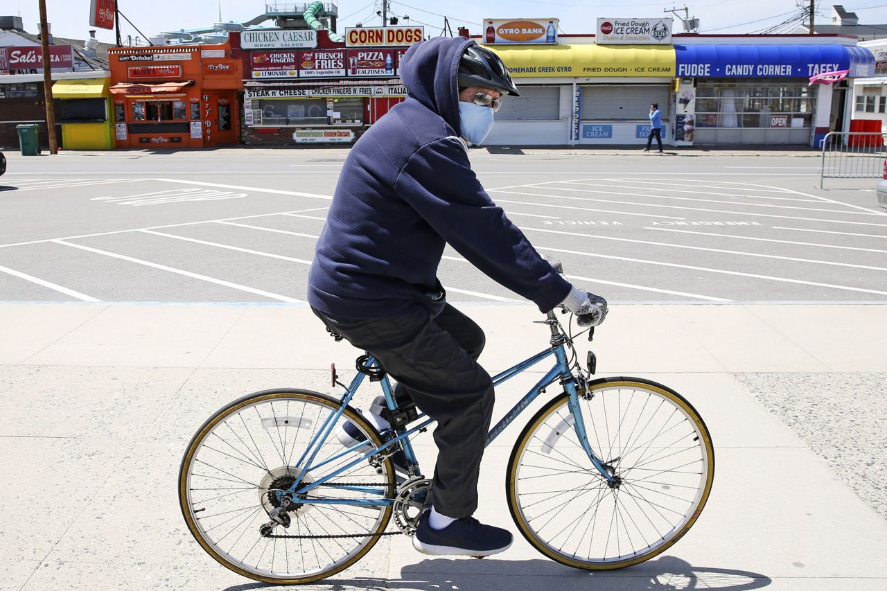 A man wearing a protective mask and gloves cycles past the Boardwalk across from the Hampton Beach State Park in Hampton, New Hampshire, on Tuesday, May 19.