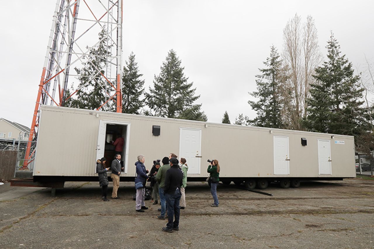 Barbara Ramey, third from left, deputy communications manager for the King County Dept. of Executive Services, briefs reporters, Tuesday, March 3, at the site in South Seattle where King County will be placing several temporary housing units like the one shown to house patients undergoing treatment and isolation.