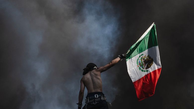 Un manifestante sostiene una bandera mexicana frente a una columna de humo proveniente de autos en llamas el 8 de junio de 2025 en el centro de Los Ángeles, California.