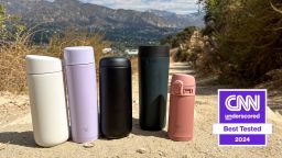 Five travel mugs placed on a dirt trail in a row, mountains in the distance behind them.