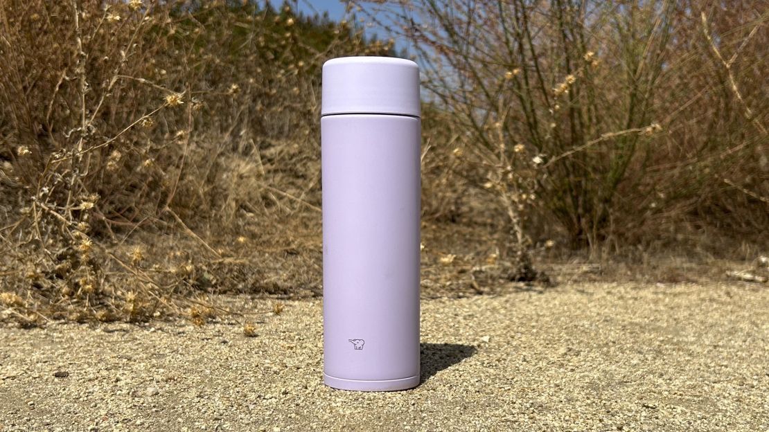 Purple travel mug placed on the ground of a hiking trail in front of brush.