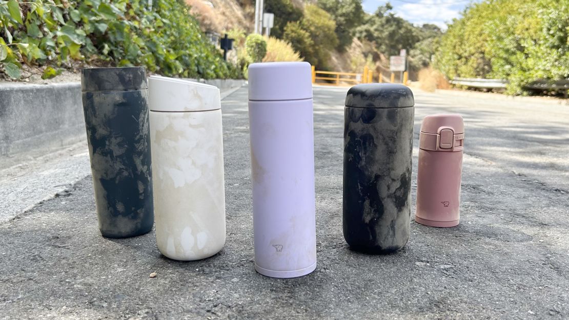 Five travel mugs arranged in arrow display, placed on a road in front of a hiking trail.