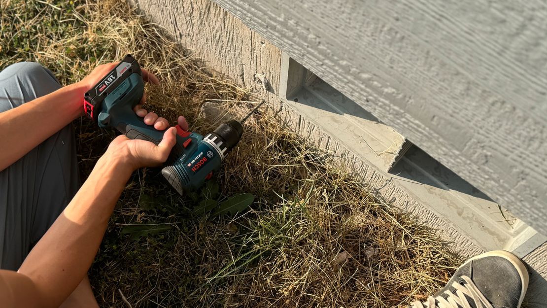 Person's arm holding a drill, using it on the side of a wooden wall. 