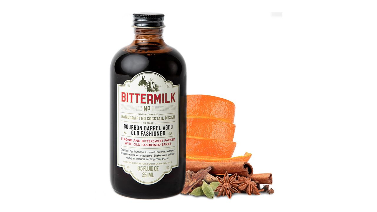 A bottle of Bittermilk Mixers Bourbon Barrel Aged Old Fashioned with several orange peels and spices on a white background.
