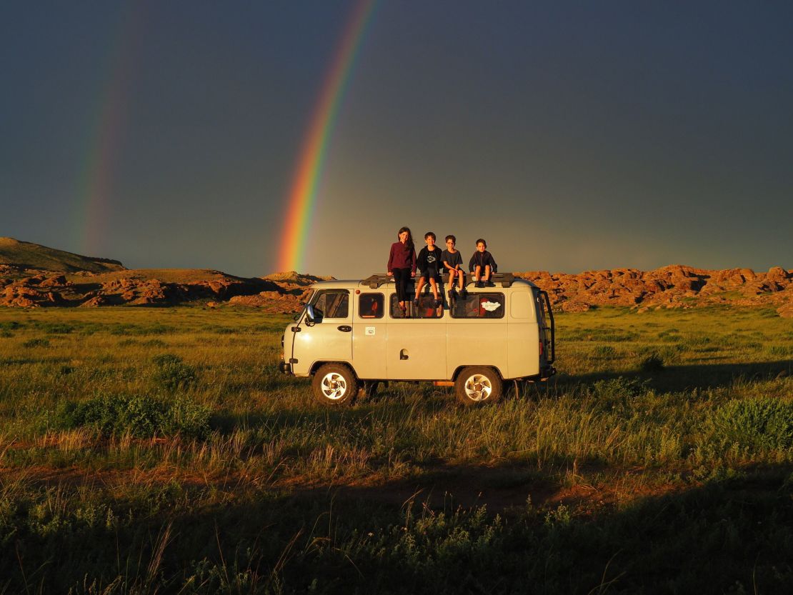Mia, Leo, Colin, and Laurent Pelletier pose on top of their camper van in front of a double rainbow while in Mongolia, in