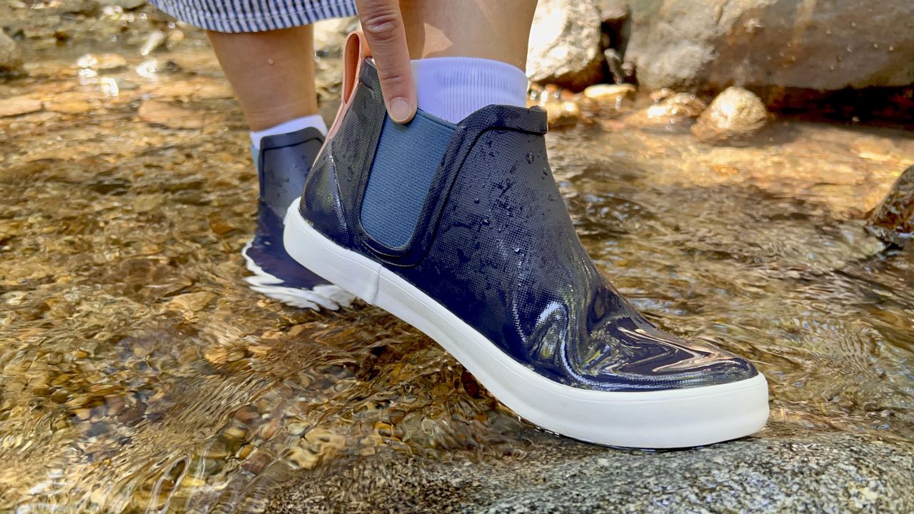 Close up of ankle rain boots stepping in a river, hand reaching down to touch the top of the boot.