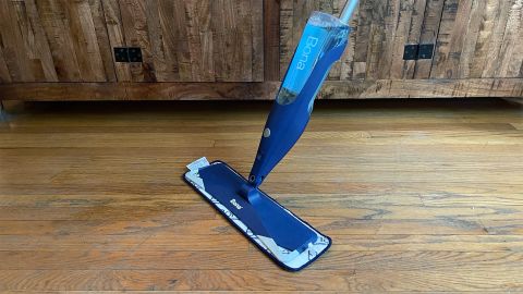 The Bona Hardwood Floor Premium Spray Mop, seen in front of a wooden cabinet scrubbing a hardwood floor.