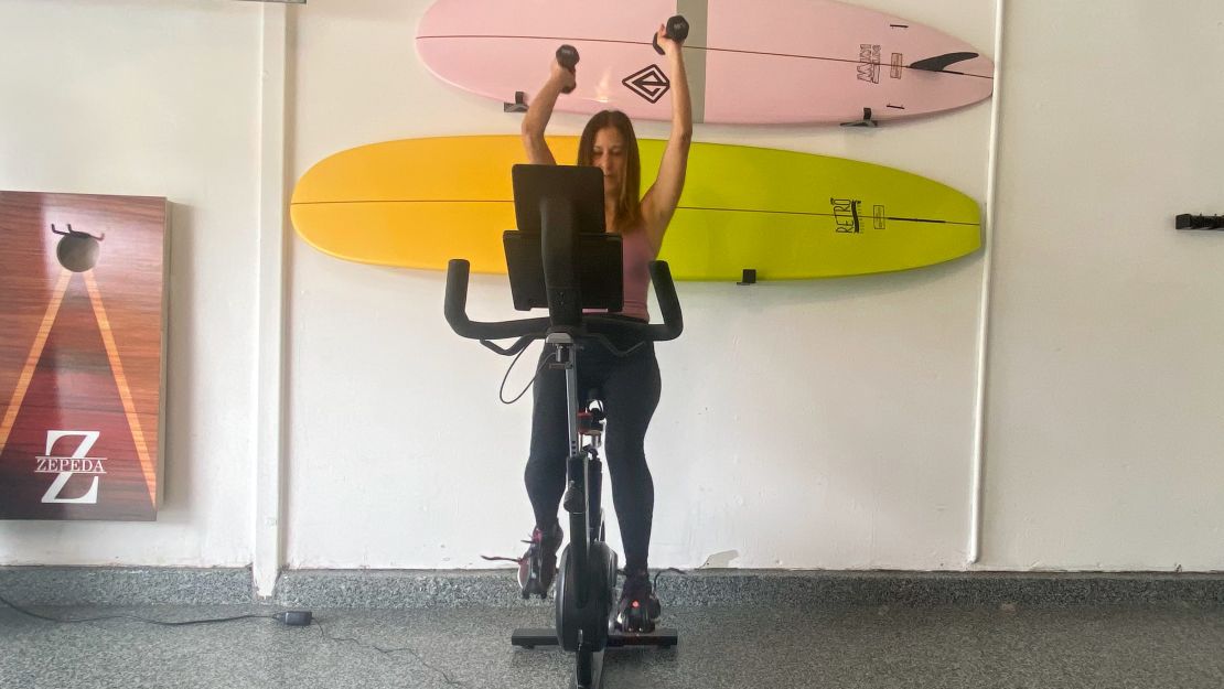 A woman cycles on the Bowflex IC Bike SE while holding dumbbells above her head.