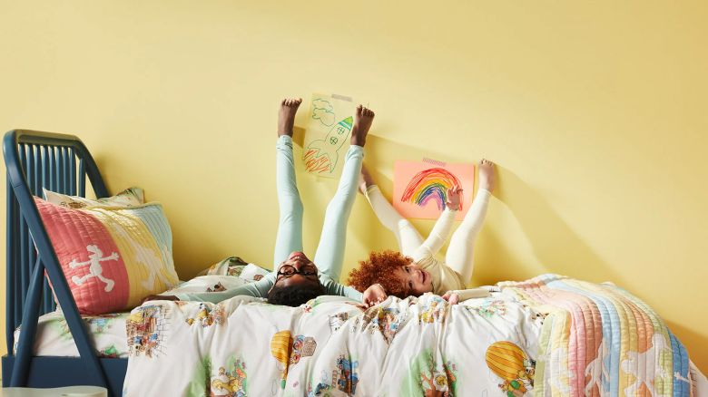 Two children have their feet on the wall as they sit on a bed fitted with Sesame Street sheets made by Brooklinen.
