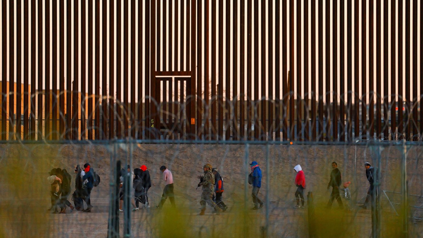 Migrants seeking asylum in the United States gather near the border wall after crossing a razor wire fence as a member of the Texas National Guard escorts them on December 19, 2024.