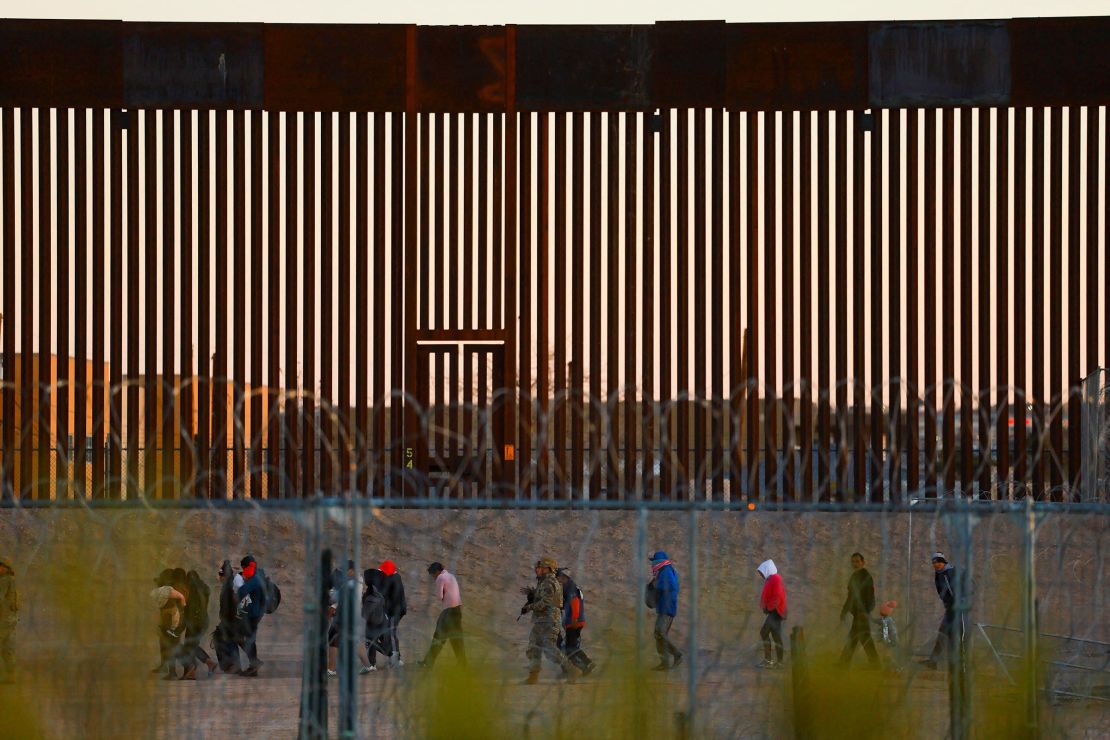 Migrants seeking asylum in the United States gather near the border wall after crossing a razor wire fence as a member of the Texas National Guard escorts them on December 19, 2024.