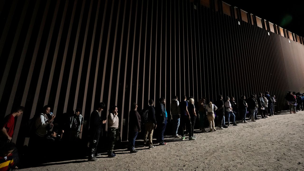 In this July 2023 photo, people line up against a border wall as they wait to apply for asylum after crossing the border from Mexico near Yuma, Arizona.