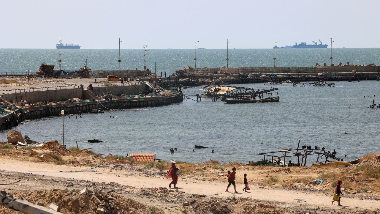 Palestinians walk past a jetty in Gaza City with a view of navy vessels off the coast as part of a humanitarian "maritime corridor" announced by US Central Command on May 17. The US military said aid deliveries began on May 17 via a temporary pier in Gaza aimed at ramping up emergency humanitarian assistance to the war-ravaged Palestinian territory.