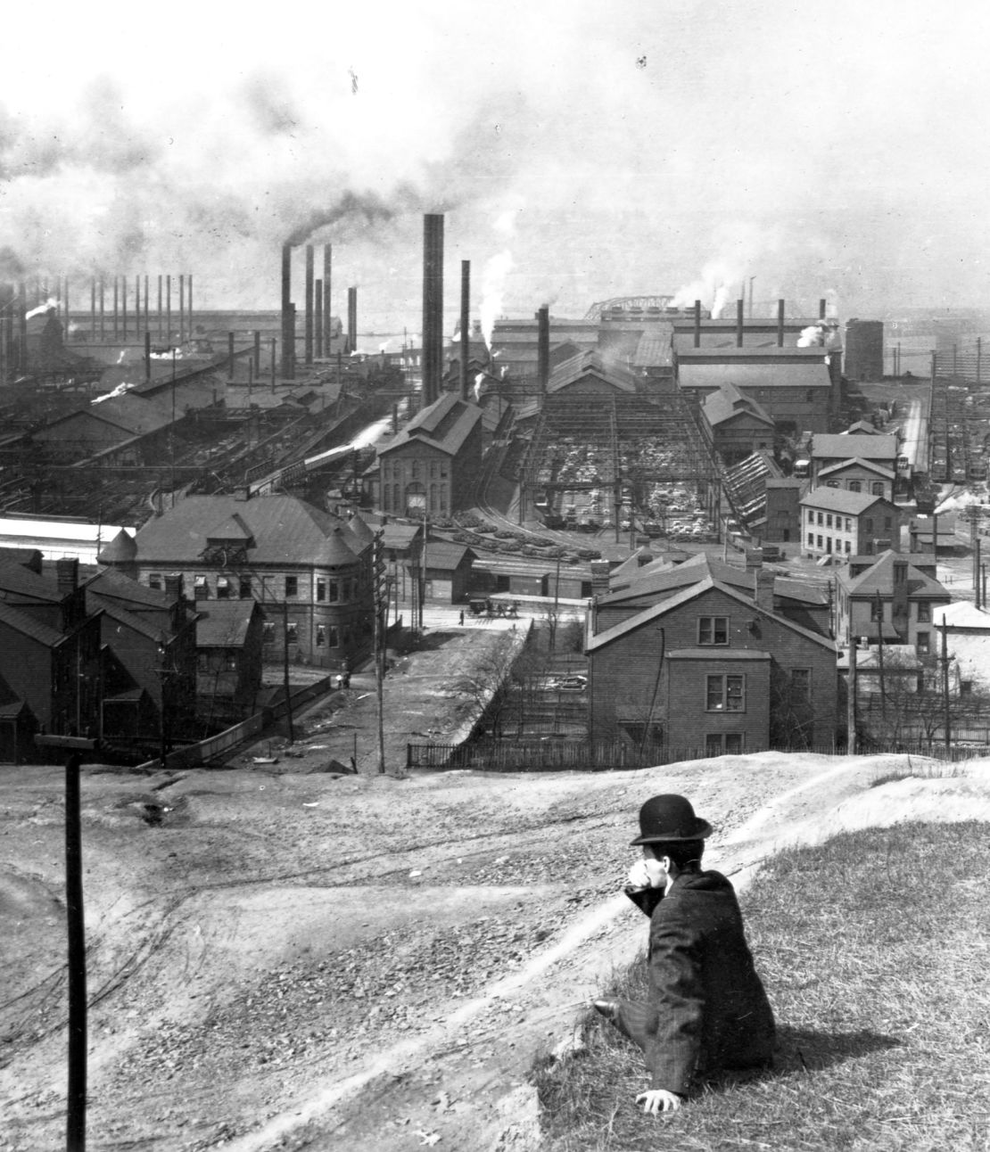 A man sits on a hill overlooking a large steel mill in Homestead, Pennsylvania in 1907.