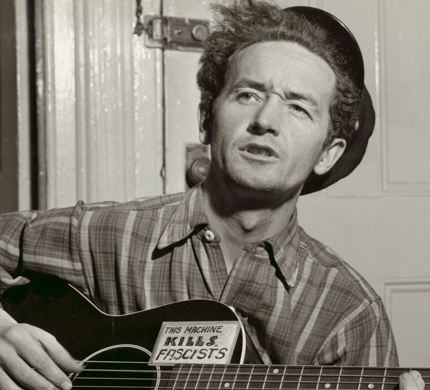 Woody Guthrie in 1943, holding his guitar with a sticker that reads, This Machine Kills Fascists.