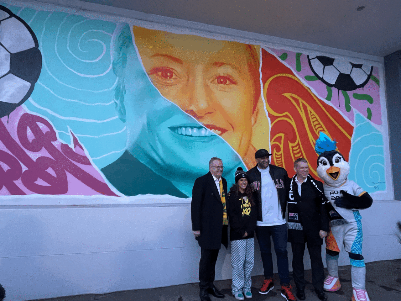 Albanese, Te Waiotu, Hoete and Hipkins pose with Tazuni, the 2023 Women's World Cup mascot, in front of the mural.