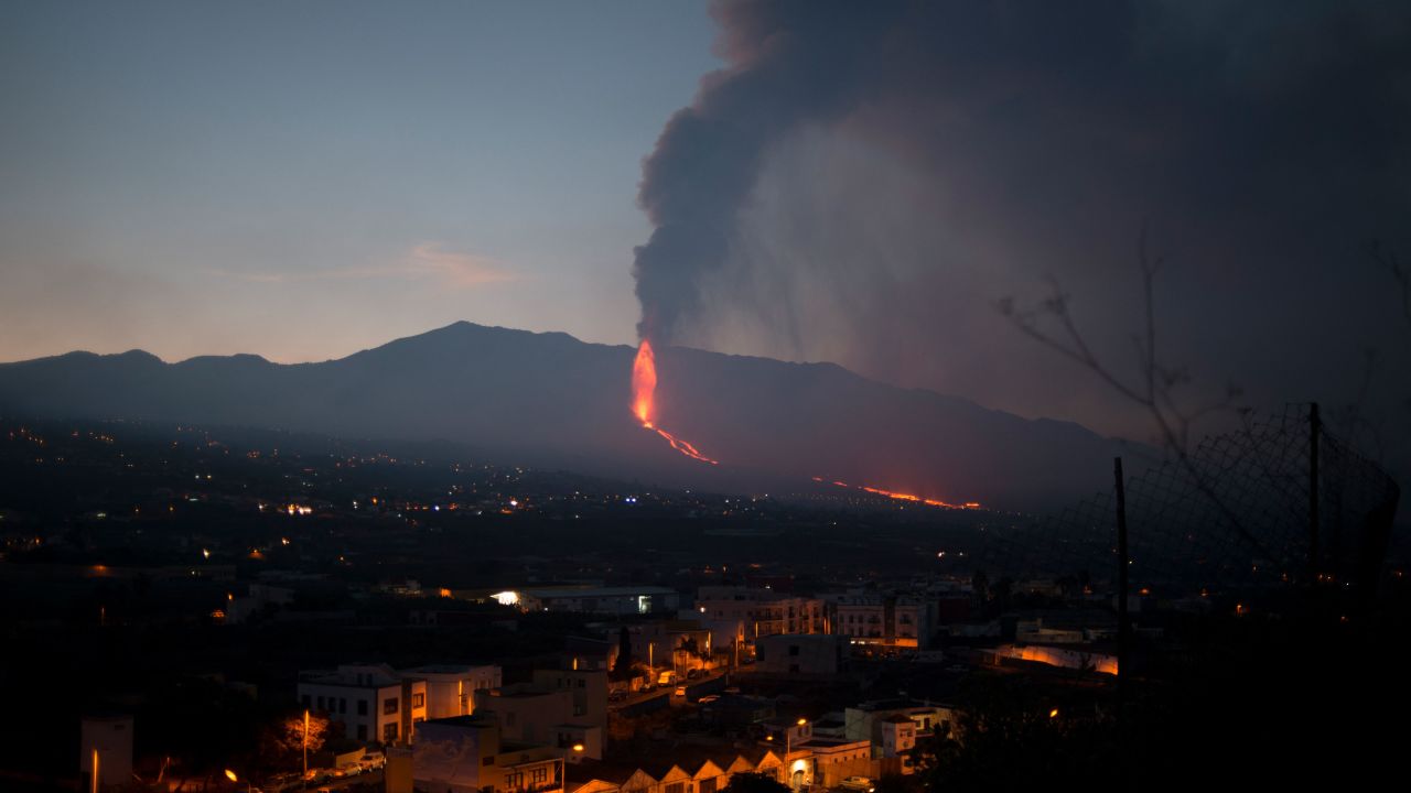 TOPSHOT - Buildings and housing are pictured as the Cumbre Vieja volcano in the background spews lava, ash and smoke, in Los Llanos de Aridane, in the Canary Island of La Palma on October 4, 2021. - A new flow of highly liquid lava emerged from the volcano erupting in Spain's Canary islands on October 1, authorities said, as a huge magma shelf continues to build on the Atlantic ocean. (Photo by JORGE GUERRERO / AFP)