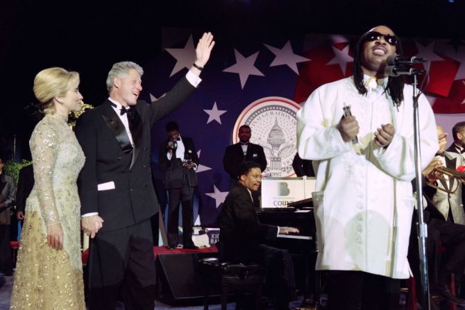 El presidente de Estados Unidos Bill Clinton flanqueado por la primera dama Hillary Rodham Clinton saluda a la multitud mientras el cantante Stevie Wonder actúa durante un baile inaugural en Washington DC el 20 de enero de 1997.