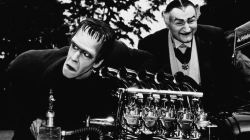 Actors Fred Gwynne (left) and Al Lewis look at a dragster car engine in a still from the television series, 'The Munsters,' circa 1964.