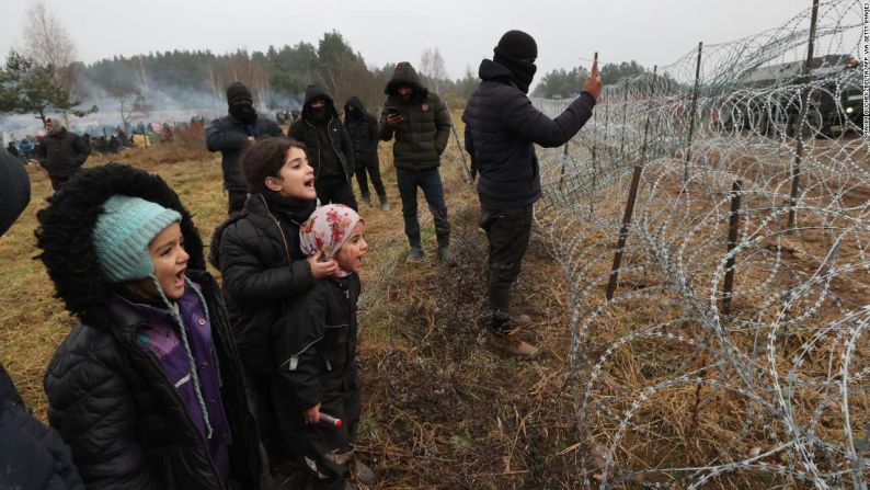 Migrantes, incluidos niños, que intentan ingresar a la UE gritan a través de la valla de alambre de púas que colocaron las autoridades polacas en la frontera con Belarús en noviembre de 2021. El presidente bielorruso, Alexander Lukashenko, ha sido acusado por la UE de orquestar la crisis migratoria tras las sanciones impuestas, pero él lo ha negado. (MAXIM GUCHEK/BELTA/AFP via Getty Images) →