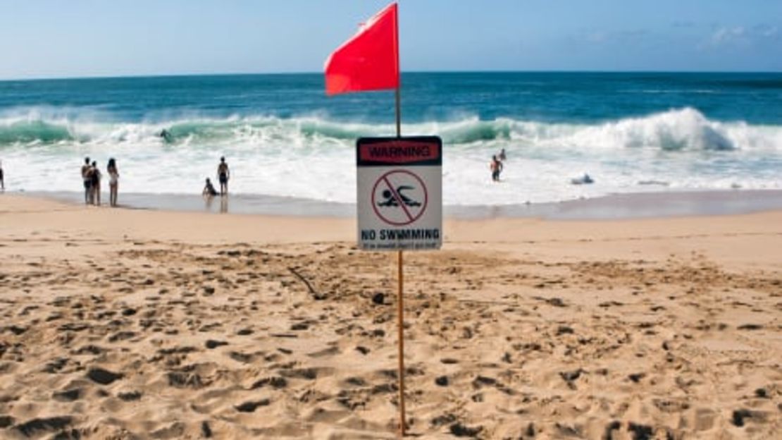 La gente nada en las olas en Waimea Bay Beach Park en la costa norte de Oahu, Hawái, bajo una bandera de advertencia. Los expertos dicen que no subestimes el poder de las olas, incluso si no hay una advertencia oficial.