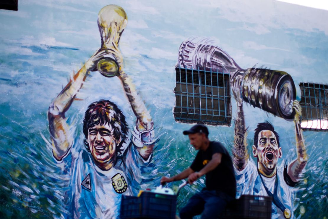 Un hombre monta su bicicleta junto a un mural que representa a Diego Armando Maradona sosteniendo la copa del mundo y a Lionel Messi sosteniendo la Copa América en Rafael Calzada el 21 de octubre de 2021 en Buenos Aires, Argentina.