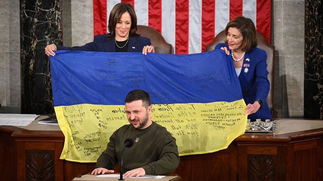 El presidente ucraniano, Volodymyr Zelensky, habla al Congreso de EE.UU., mientras la vicepresidenta, Kamala Harris, y la presidenta de la Cámara, Nancy Pelosi, sostienen una bandera ucraniana firmada por los soldados en el frente de batalla.