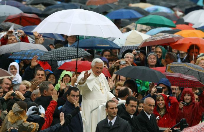 Benedicto saluda desde debajo de un paraguas cuando llega para dirigir su audiencia general semanal en la Plaza de San Pedro en el Vaticano en octubre de 2007.