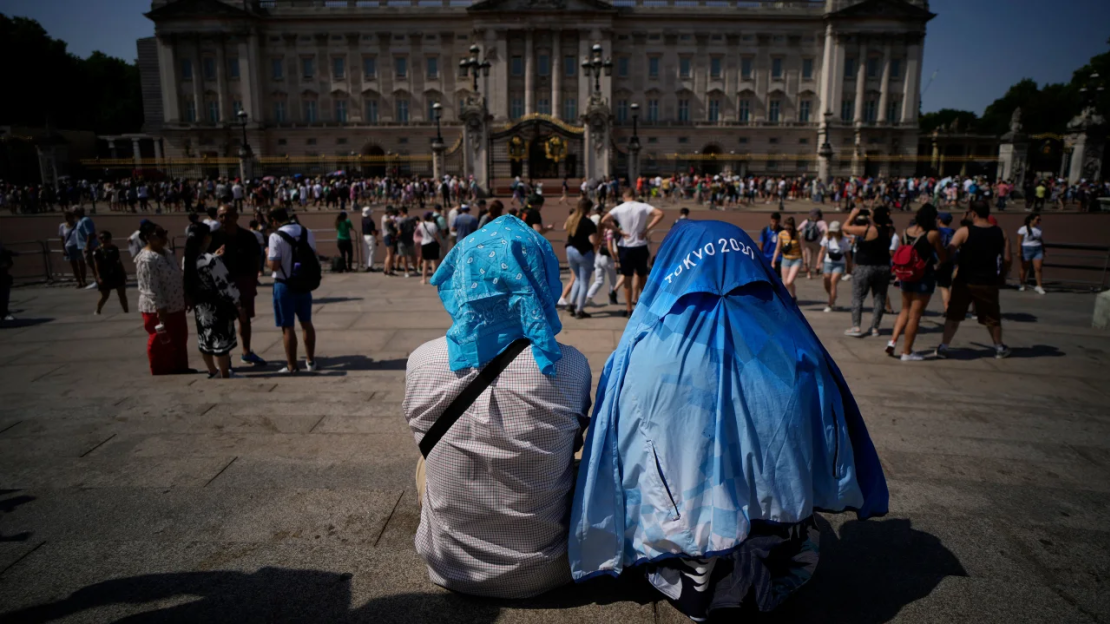 La gente se sienta cubriendo sus cabezas del sol después de una versión reducida de la ceremonia del Cambio de Guardia que tuvo lugar fuera del Palacio de Buckingham, durante el clima caluroso en Londres, el lunes 18 de julio de 2022.