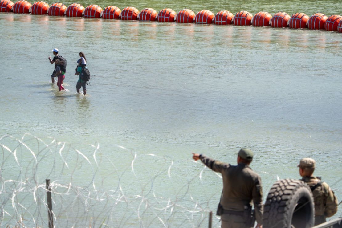 Agentes de la patrulla de carreteras del Departamento de Seguridad Pública de Texas (DPS, por sus siglas en inglés) miran por encima del río Grande mientras los migrantes caminan junto a una cadena de boyas colocadas en el agua a lo largo de la frontera con México en Eagle Pass, Texas, el 15 de julio de 2023, para impedir la entrada ilegal de migrantes a EE.UU.