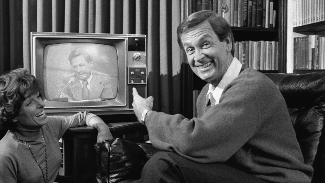 El presentador de un programa de juegos estadounidense Bob Barker se señala a sí mismo en una pantalla de televisión cercana mientras su esposa Dorothy Jo Barker (1924 - 1981) mira y ríe, el 4 de noviembre de 1977. Crédito: Imágenes CBS/Getty