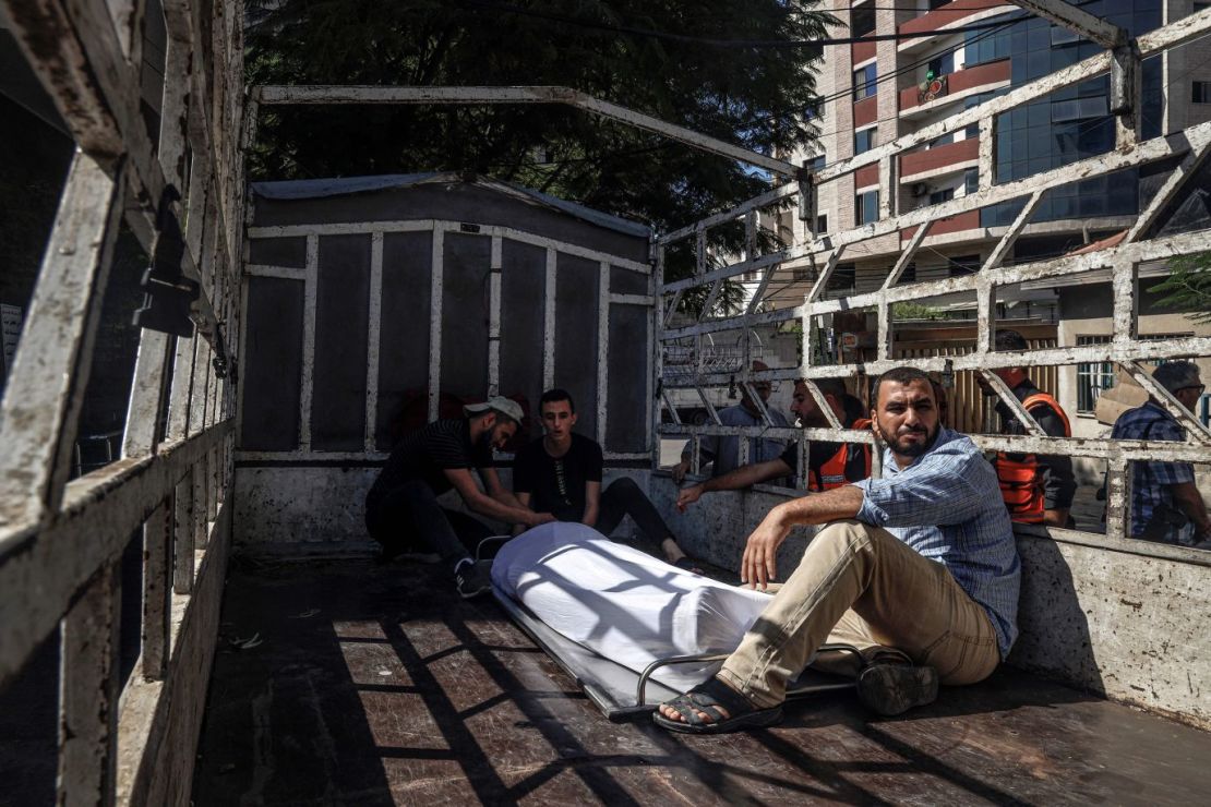 Afuera de un hospital en Gaza, hombres sentados junto al cuerpo cubierto de un militante palestino muerto durante los enfrentamientos del sábado 7 de octubre.