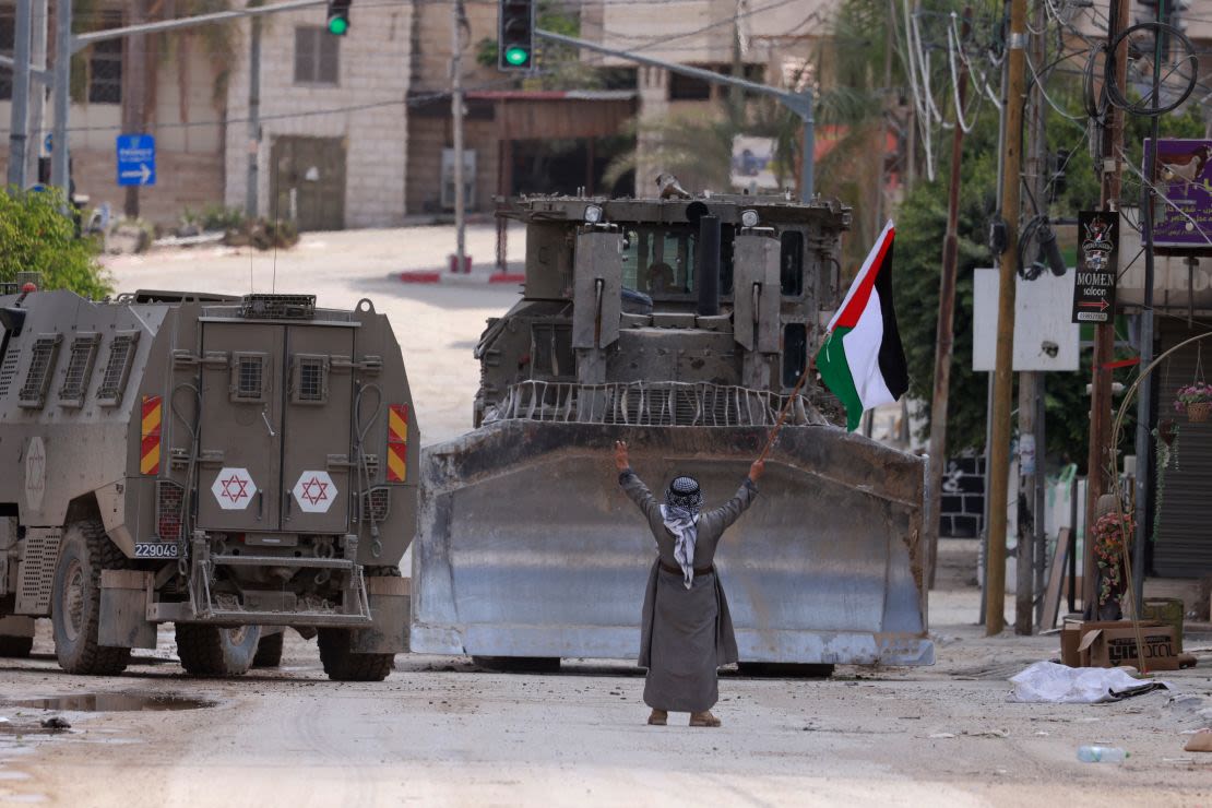 Un palestino ondea una bandera nacional mientras vehículos blindados israelíes circulan por una calle durante una redada en Tulkarem el martes. Crédito: JAAFAR ASHTIYEH/AFP/AFP vía Getty Images