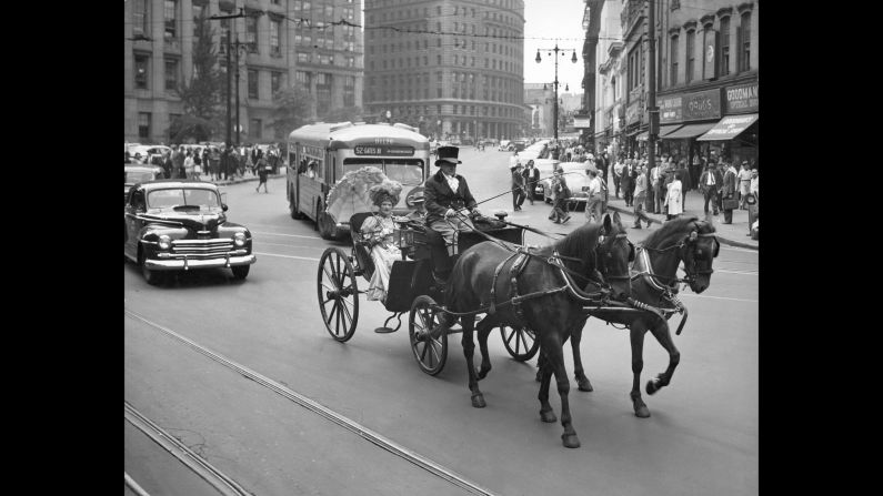 Un conductor de carruaje con sombrero de copa y una pasajera con un vestido con mangas de pierna de cordero que se protege con un elegante paraguas evocan espléndidamente el pasado en una calle de la ciudad de Nueva York en 1947.