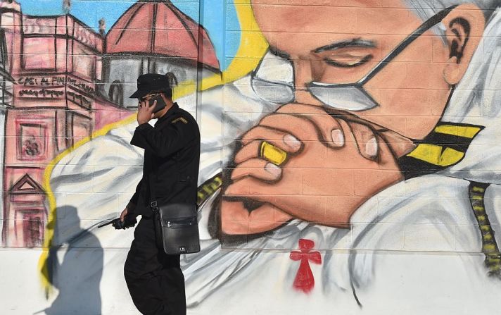 Ecatepec, México: Un miembro de seguridad habla en su teléfono móvil junto a una pintura en un mural que representa a Francisco en Ecatepec.