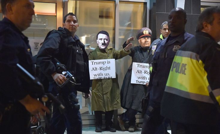 Dos hombres con máscaras de Adolf Hitler y Benito Mussolini protestan en Washington haciendo el saludo nazi con carteles que dicen “Trump está de bien nosotros”, mientras policías vigilan la seguridad del lugar.