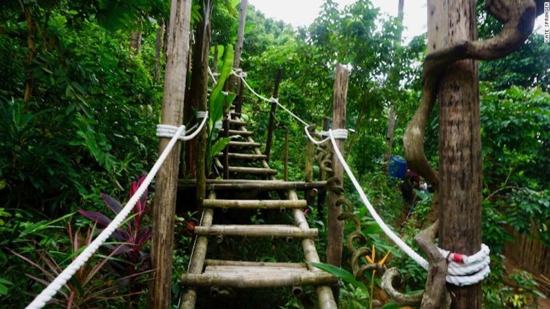 Caminata por el bosque — Sigue las señales por un sendero a través del bosque. Una escalera conduce a los viajeros por una ladera exuberante desde donde se pueden ver unas tiendas contemporáneas entre las ramas de los árboles.